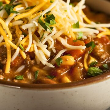 Close-up of Southwest beef and corn chili in a speckled ceramic bowl, topped with shredded cheddar and Monterey Jack cheese and chopped green onions. Thick tomato-based chili with ground beef, sweet corn, and beans visible in a rich broth. Cozy high protein chili recipe, hearty one pot dinner.