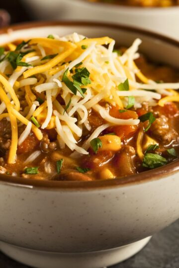 Close-up of Southwest beef and corn chili in a speckled ceramic bowl, topped with shredded cheddar and Monterey Jack cheese and chopped green onions. Thick tomato-based chili with ground beef, sweet corn, and beans visible in a rich broth. Cozy high protein chili recipe, hearty one pot dinner.
