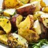 Close up of oven baked red potatoes with Parmesan and thyme, cut into wedges with crispy golden centers and red skins. Roasted garlic cloves and fresh rosemary sprigs are scattered over the potatoes on a white plate, highlighting the browned edges and herb seasoning.