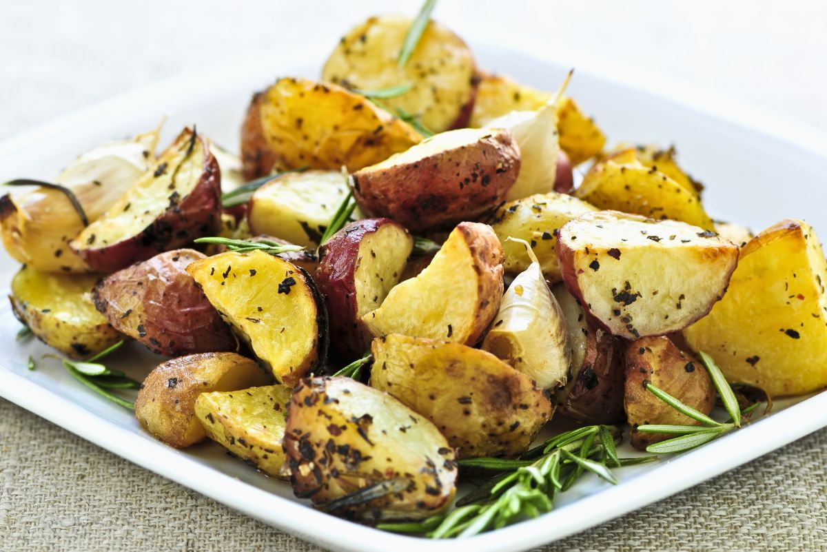 Oven baked red potatoes with Parmesan and thyme on a white square plate, cut into wedges with crispy golden centers and browned red skins. Roasted garlic cloves and fresh rosemary sprigs are scattered around the potatoes, highlighting the herb seasoned, crispy edges.