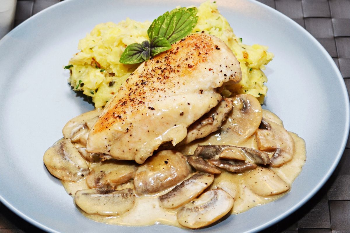 Skillet herb chicken breast with golden sear, topped with creamy lemon mushroom sauce and sliced mushrooms on a gray plate. Served with mashed potatoes and fresh basil garnish. High protein chicken dinner with rich mushroom gravy.