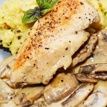 Close-up of skillet herb chicken breast with golden seared top, covered in creamy lemon mushroom sauce and sliced mushrooms. Served on a gray plate with mashed potatoes and fresh basil garnish. High protein chicken dinner with rich mushroom gravy.