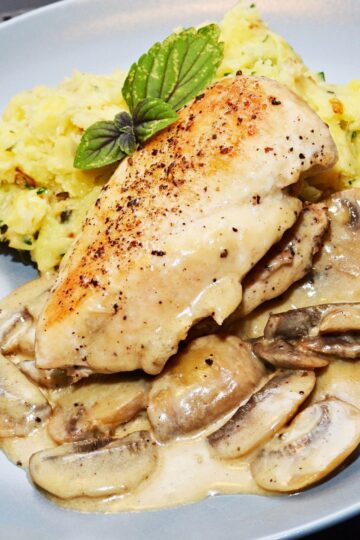 Close-up of skillet herb chicken breast with golden seared top, covered in creamy lemon mushroom sauce and sliced mushrooms. Served on a gray plate with mashed potatoes and fresh basil garnish. High protein chicken dinner with rich mushroom gravy.