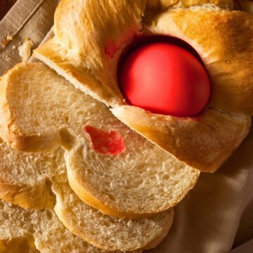 Close-up of traditional Greek Tsoureki Easter bread with a glossy braided crust and a bright red dyed egg baked into the center. Several soft slices show the fluffy, slightly sweet interior. Classic homemade Greek Easter bread on a linen cloth with a knife nearby.