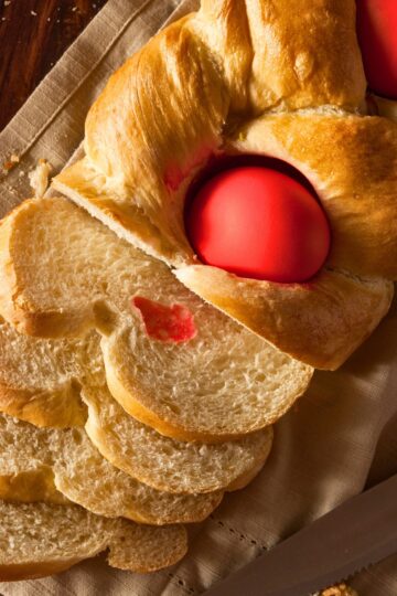 Close-up of traditional Greek Tsoureki Easter bread with a glossy braided crust and a bright red dyed egg baked into the center. Several soft slices show the fluffy, slightly sweet interior. Classic homemade Greek Easter bread on a linen cloth with a knife nearby.