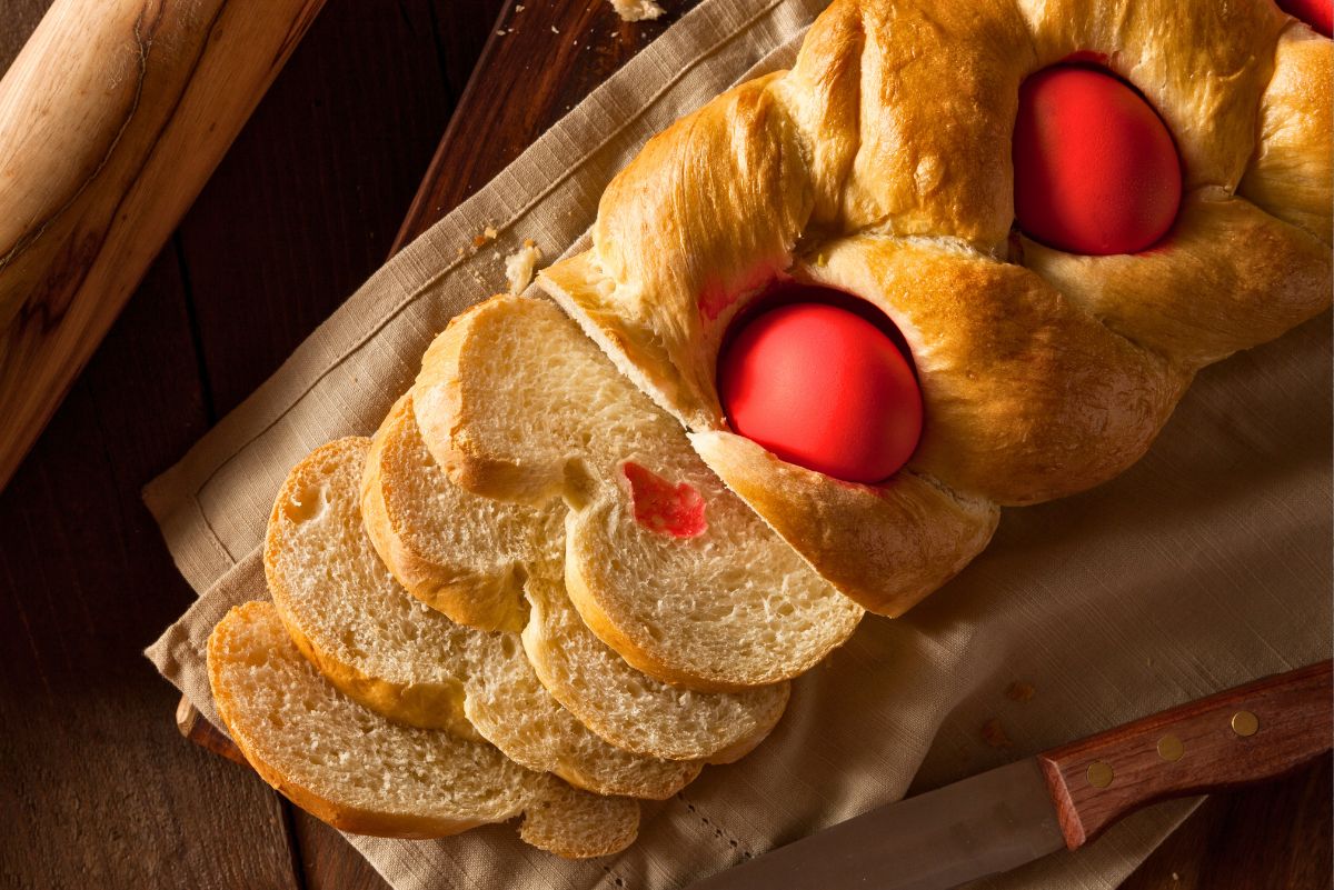 Braided traditional Greek Tsoureki Easter bread with glossy golden crust and bright red dyed eggs baked into the dough. Several soft slices are fanned out, showing the fluffy interior. Bread rests on a linen cloth over a wooden table with a rolling pin and knife nearby. Classic homemade Greek Easter bread.