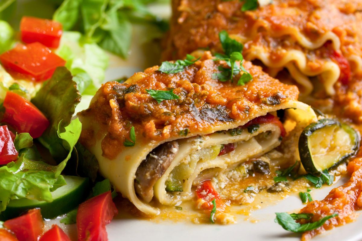 Plate of vegetarian lasagna roll ups filled with mushrooms, spinach, zucchini, and red bell pepper, topped with marinara sauce, melted cheese, and fresh parsley. Served with a side salad of lettuce, cucumber, and tomato. High protein meatless Italian pasta dinner recipe.