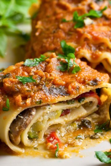 Close-up of vegetarian lasagna roll ups filled with mushrooms, spinach, zucchini, and red bell pepper, topped with rich marinara sauce, melted cheese, and fresh parsley. Creamy ricotta-style filling wrapped in tender pasta sheets on a white plate. High protein meatless Italian dinner recipe.