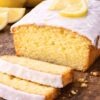 Iced lemon pound cake loaf with thick white glaze on a rustic wooden board, topped with a lemon slice. Two uneven slices are cut in front, showing the moist yellow crumb, with a few crumbs scattered around. Whole lemons and lemon wedges sit softly blurred in the background.