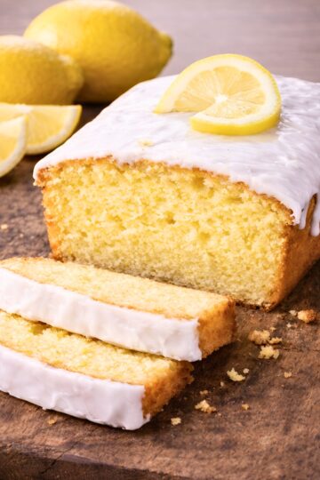 Iced lemon pound cake loaf with thick white glaze on a rustic wooden board, topped with a lemon slice. Two uneven slices are cut in front, showing the moist yellow crumb, with a few crumbs scattered around. Whole lemons and lemon wedges sit softly blurred in the background.
