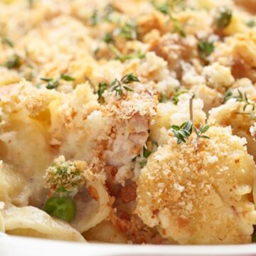 Close-up tuna noodle casserole in a red baking dish with creamy noodles, peas, tuna, herbs, and a golden potato chip crumb topping. Baked comfort food casserole with a crisp crunchy top and rich creamy texture.