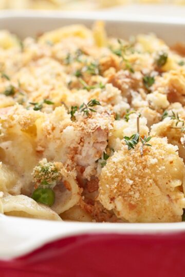 Close-up tuna noodle casserole in a red baking dish with creamy noodles, peas, tuna, herbs, and a golden potato chip crumb topping. Baked comfort food casserole with a crisp crunchy top and rich creamy texture.