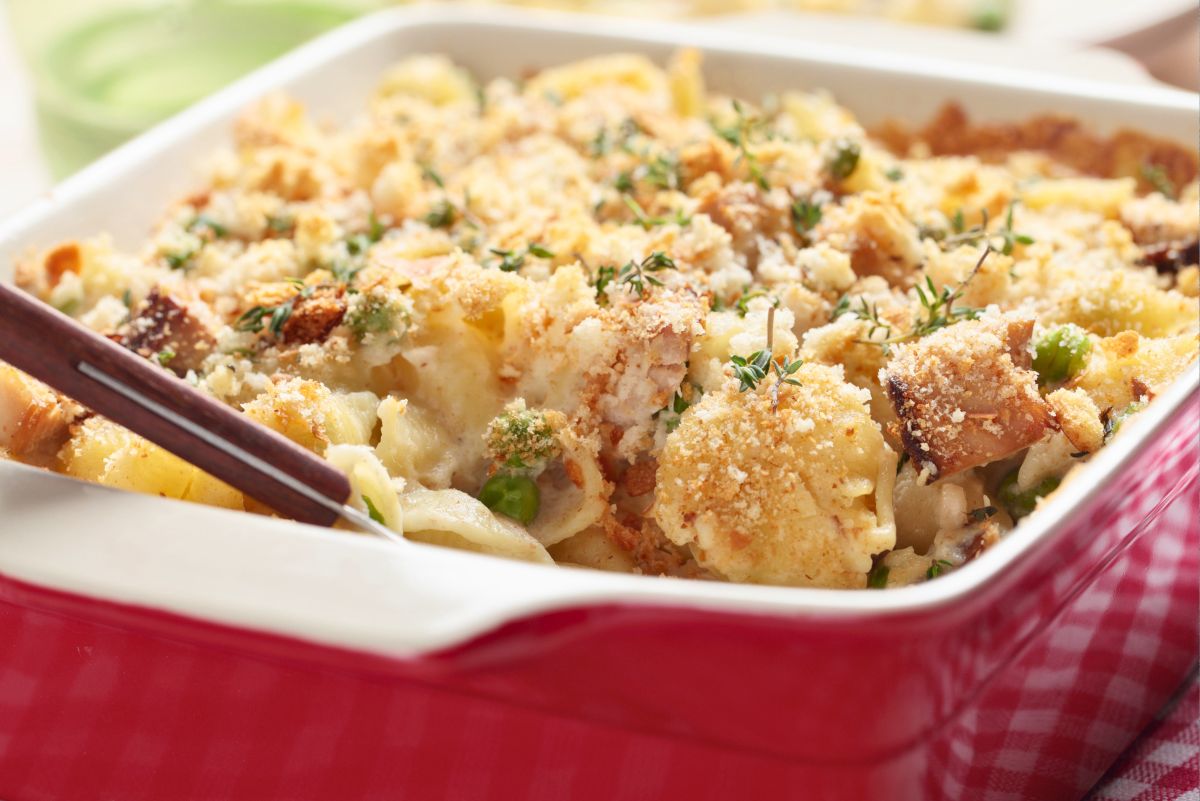 Tuna noodle casserole with potato chip topping in a red baking dish. Creamy noodles, tuna, peas, herbs, and golden crunchy crumbs fill the casserole, with a serving spoon at the side. Classic comfort food casserole in a close-up food photo.