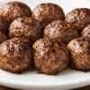 Close-up of homemade baked beef meatballs on a white plate over a rustic wood table. The Italian meatballs are browned on the outside with a juicy, glossy finish.