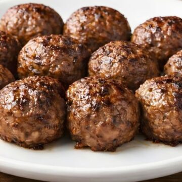 Close-up of homemade baked beef meatballs on a white plate over a rustic wood table. The Italian meatballs are browned on the outside with a juicy, glossy finish.