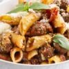 Close-up of rigatoni pasta with Italian sausage in tomato basil sauce, topped with grated Parmesan and fresh basil in a white bowl. Rustic pasta dinner with chunky sausage, tomato pieces, and ridged rigatoni on a light background.