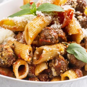 Close-up of rigatoni pasta with Italian sausage in tomato basil sauce, topped with grated Parmesan and fresh basil in a white bowl. Rustic pasta dinner with chunky sausage, tomato pieces, and ridged rigatoni on a light background.