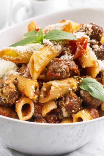 Close-up of rigatoni pasta with Italian sausage in tomato basil sauce, topped with grated Parmesan and fresh basil in a white bowl. Rustic pasta dinner with chunky sausage, tomato pieces, and ridged rigatoni on a light background.