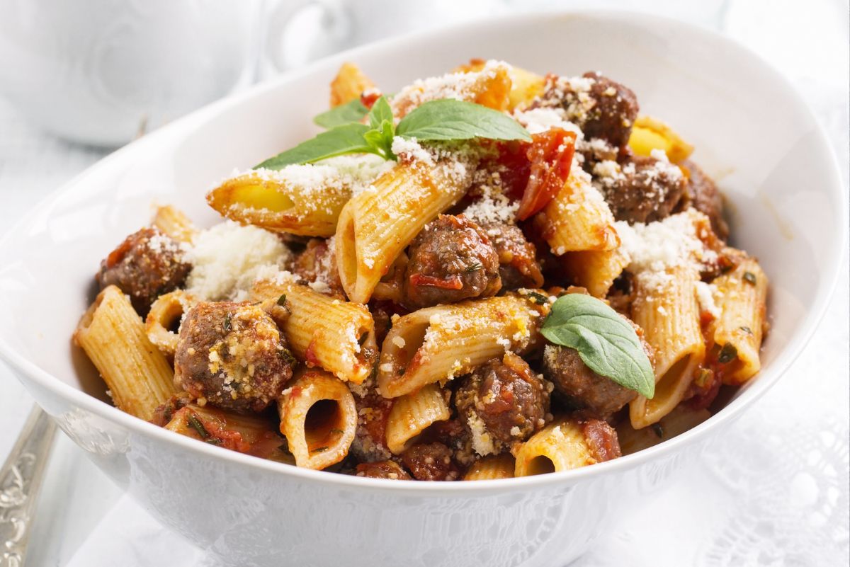 White bowl of rigatoni with Italian sausage in tomato basil sauce, topped with grated Parmesan and fresh basil leaves. Close-up pasta dinner with browned sausage pieces, tomato chunks, and ridged rigatoni on a light table setting.