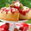 Upside Down Strawberry Marshmallow Cake on a white cake stand with fresh strawberries, a daisy garnish, and a slice cut out. A red plate, gingham napkin, and dessert fork sit below with a blurred green background.