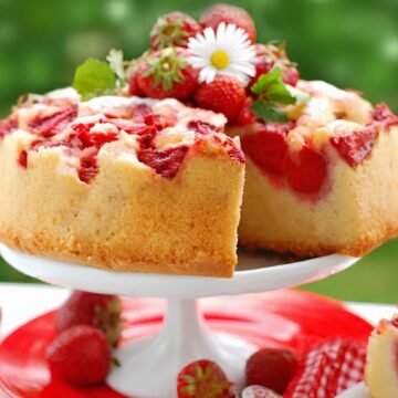 Upside Down Strawberry Marshmallow Cake on a white cake stand with fresh strawberries, a daisy garnish, and a slice cut out. A red plate, gingham napkin, and dessert fork sit below with a blurred green background.