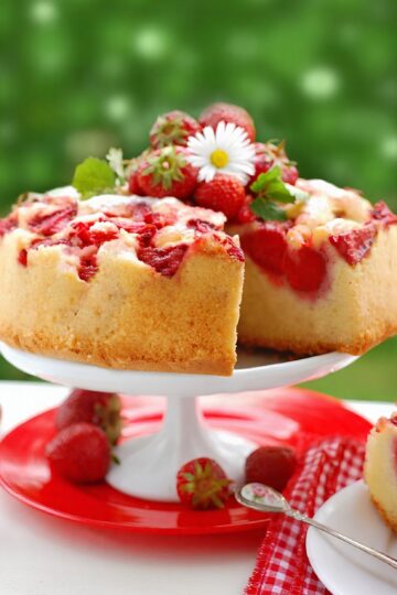 Upside Down Strawberry Marshmallow Cake on a white cake stand with fresh strawberries, a daisy garnish, and a slice cut out. A red plate, gingham napkin, and dessert fork sit below with a blurred green background.