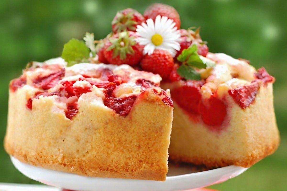 Upside Down Strawberry Marshmallow Cake on a white cake stand, topped with baked strawberries, fresh strawberries, green leaves, and a daisy garnish against a blurred green background.